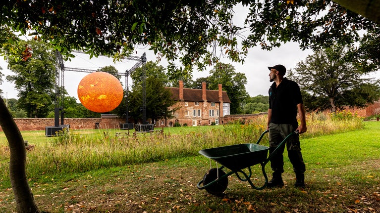 A gardener with his wheelbarrow looks over towards the bright yellow-red of Helios, the art installation of the sun, with the Charterhouse nestled on its right hand side